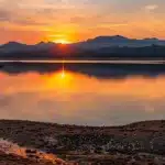a breathtaking sunset casts vibrant hues of orange and pink across a serene lake, surrounded by majestic mountains reflecting in the still water.