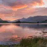 a breathtaking sunset casts vibrant hues of orange and pink across a tranquil lake, with silhouetted mountains reflecting on its glassy surface, evoking a sense of peaceful serenity.