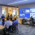a dynamic urban business setting featuring a sleek conference room with a large digital display, where professionals engage in animated discussions, illuminated by modern overhead lighting.