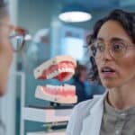 a close-up of a thoughtful speech therapist in a sleek, modern office, demonstrating clear enunciation techniques, with a detailed anatomical model of a mouth and teeth prominently displayed in the background, emphasizing the connection between oral health and communication clarity.