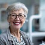 a confident 72-year-old woman, smiling brightly in a modern dental clinic, showcases her newly fitted dentures against a sleek, vibrant backdrop of dental equipment, emphasizing the theme of renewed confidence and aging gracefully.