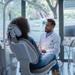a modern dental office features a young adult thoughtfully consulting with a dentist, surrounded by high-tech equipment and informative dental charts, highlighting the pros and cons of getting dentures in one's 20s.