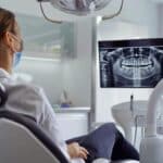 a modern dental clinic interior showcases a concerned patient seated in a sleek dental chair, as a focused dentist examines a digital x-ray displayed on a high-tech monitor, highlighting the complexities and potential complications of dental implants.